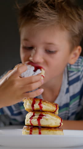 A closeup of a child eating pancakes with a spoon. A stack of pancakes with red jam is on a plate in the foreground, with the child taking a bite.