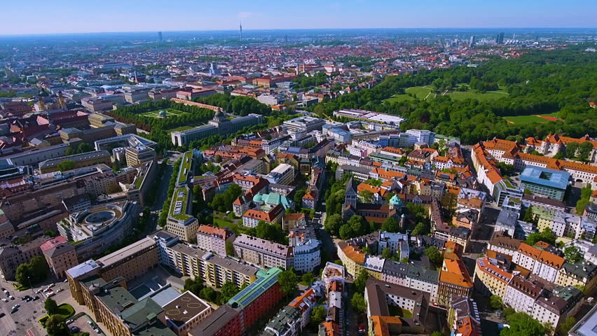 4K drone panorama of Munich with Hofgarten and the Bavarian State Chancellery, vast Englischer Garten and red-roofed Altstadt, Olympiaturm on the horizon under clear summer skies.