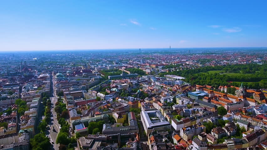 4K drone panorama of Munich on a crystal blue day: tree lined Ludwigstrabe, Frauenkirche and Theatinerkirche domes, Olympiaturm and the Englischer Garten spreading across the horizon.