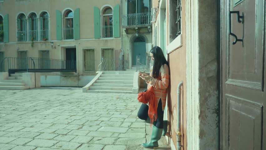 A young woman stands against a wall in a peaceful corner of Venice, surrounded by charming architecture. Her pose suggests relaxation as she checks her phone, showcasing the blend of modern life in a 