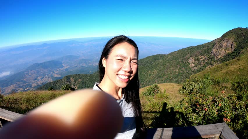 A cheerful young woman takes a selfie against a stunning mountain backdrop under a clear blue sky. The image captures her happiness and the picturesque landscape, inviting viewers to explore the beaut