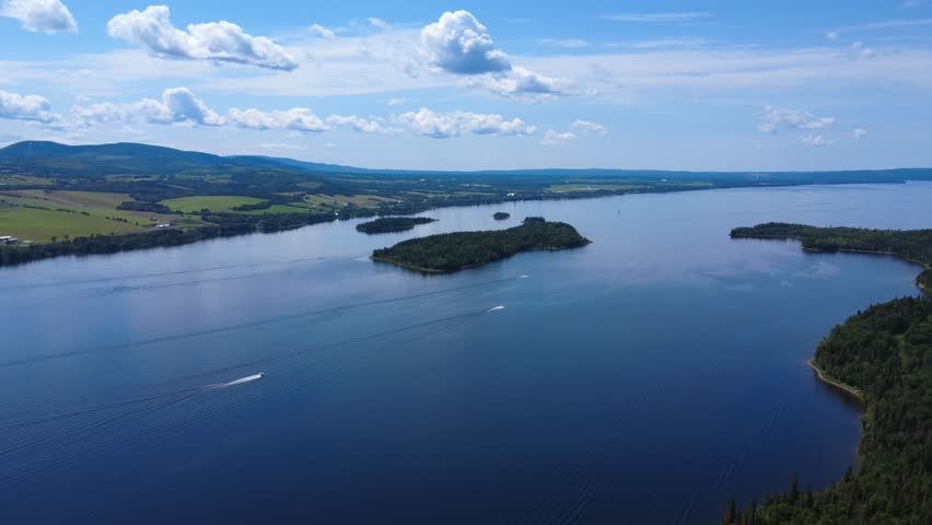 Panoramic aerial view of a group of pleasure boats all heading in the same direction near an island emerging from a vast freshwater lake along a mountainous agricultural valley under a cloudy sky. 