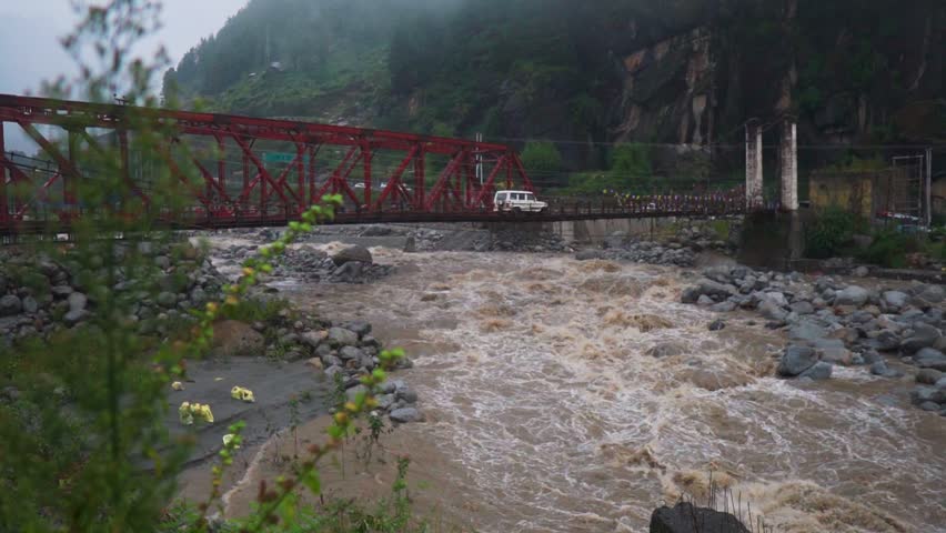 Brown monsoon waters surge under a metal bridge with crossing vehicles in Manali, Himachal Pradesh, India. Lush greenery in mountains, showing seasonal flooding and river force