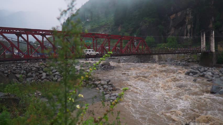 Brown monsoon waters surge under a metal bridge with crossing vehicles in Manali, Himachal Pradesh, India. Lush greenery in mountains, showing seasonal flooding and river force