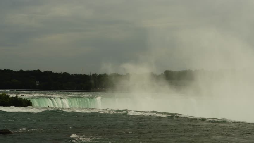 A wide shot of Niagara Falls as mist rises above the cascading water under an overcast sky. Gentle waves move across the river