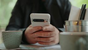 Close-up of a person using a smartphone at a cafe table, with a coffee cup and tin of utensils. Male hands hold smartphone and send message while sitting at restaurant table - Powered by Shutterstock - Get 15% off with code: PIKWIZARD15
