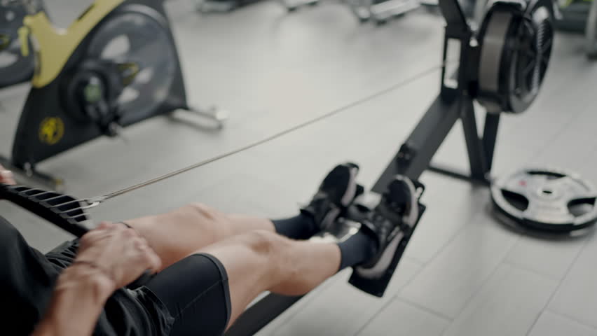 Focused young male in black athletic gear using a rowing machine at a well-equipped gym, highlighting fitness and determination. Young Man Exercising on a Rowing Machine in Modern Gym