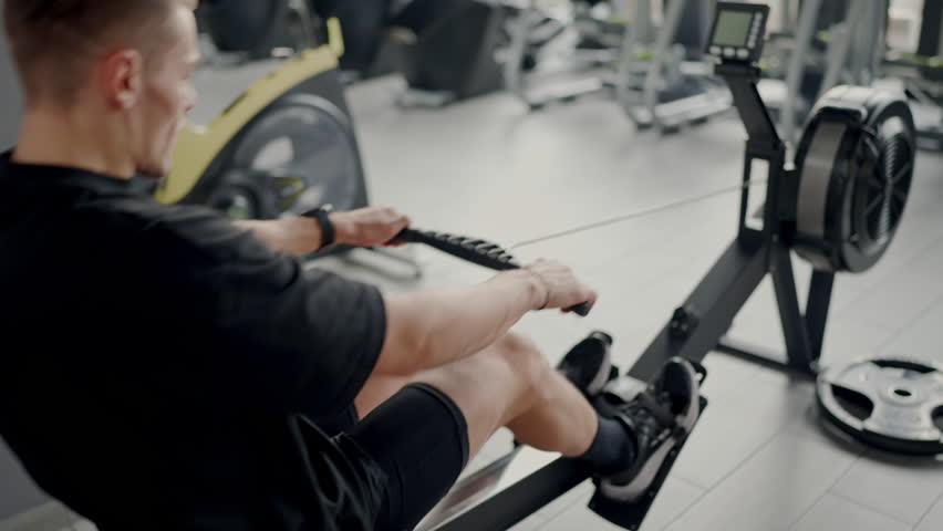 Focused young male in black athletic gear using a rowing machine at a well-equipped gym, highlighting fitness and determination. Young Man Exercising on a Rowing Machine in Modern Gym