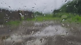 A close-up view through a rain-streaked window, blurring the world outside. Prominent raindrops create an abstract pattern on the glass, partially obscuring a road and lush green foliage. - Powered by Shutterstock - Get 15% off with code: PIKWIZARD15