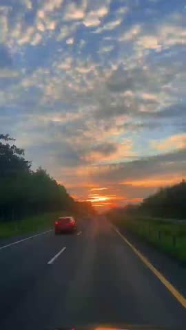 Serene sunset on a highway, with car in red driving towards the horizon. The sky is filled with beautiful altocumulus clouds and warm sunlight, capturing the feeling of travel and adventure.