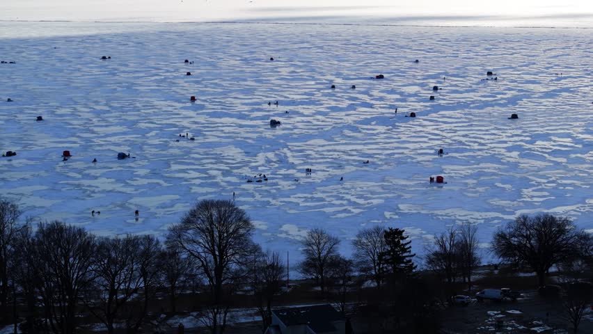 Frozen lake surface with ice fishing tents and blowing snow across textured cracked surface, drone at golden hour