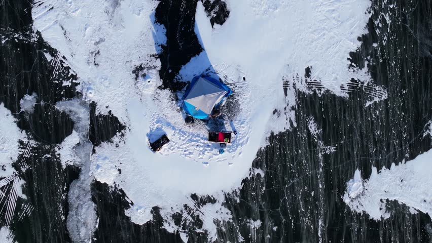 Winter ice fishing with tents and snow blowing across Lake Geneva Wisconsin, slow motion aerial top shot static