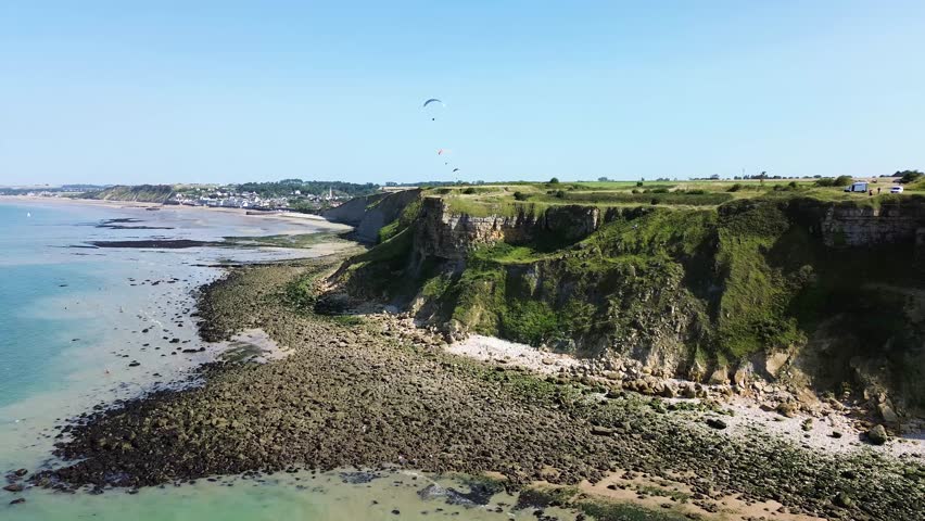 Drone footage gliding along the Arromanches cliffs, showing their majestic drop into the blue sea, framed by lush green fields under a cloudless sky.