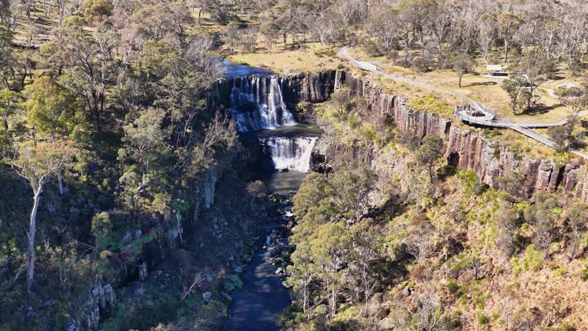 Drone camera smoothly descends above Ebor Waterfall, revealing cascading water, rugged cliffs, and dense forest in bright natural daylight