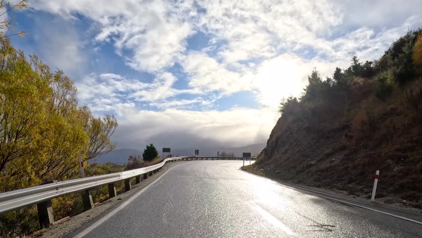 A car travels along a winding farm road through autumn hills and fields in Queenstown, New Zealand, under dramatic, partly cloudy sunlight