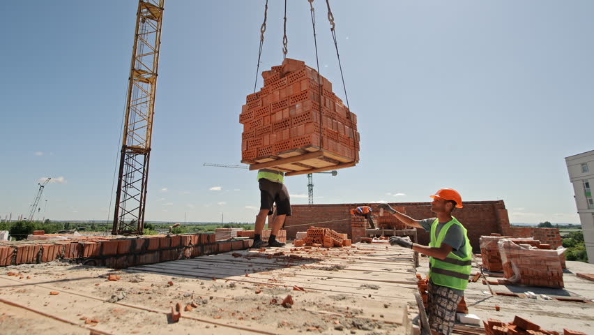Construction worker guiding brick pallet lifted by crane. Worker in safety gear directs a large pallet of hollow red bricks being lifted by crane at a building site on a sunny day.