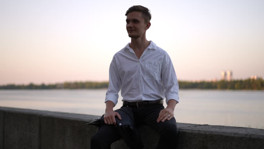 The bride in a white flowing dress leans in to kiss her groom wearing a white shirt. They hold each other closely near the water, sharing an intimate slow motion moment of tenderness and love.