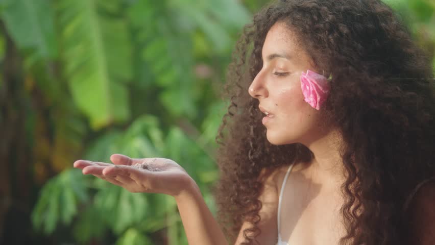 Close-up captures a woman with a pink flower tucked behind her ear, gently blowing ceremonial powder from her hand in a lush green setting, suggesting a ritual or symbolic gesture.