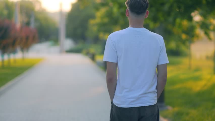 A teenage boy walks alone down a peaceful park path at sunset, hands in pockets. A quiet, emotional moment of solitude and reflection in a warm urban setting.