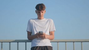 Teen boy leans against a railing and looks down at his phone with a serious face. The clear blue sky and casual vibe capture a moment of quiet modern life. - Powered by Shutterstock - Get 15% off with code: PIKWIZARD15