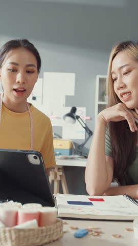 Vertical Screen : Two Asia cheerful lady fashion designer teamwork with casual on table use tablet meeting discuss final design share idea in shop. Lady tailor partner start up business cloth shop.