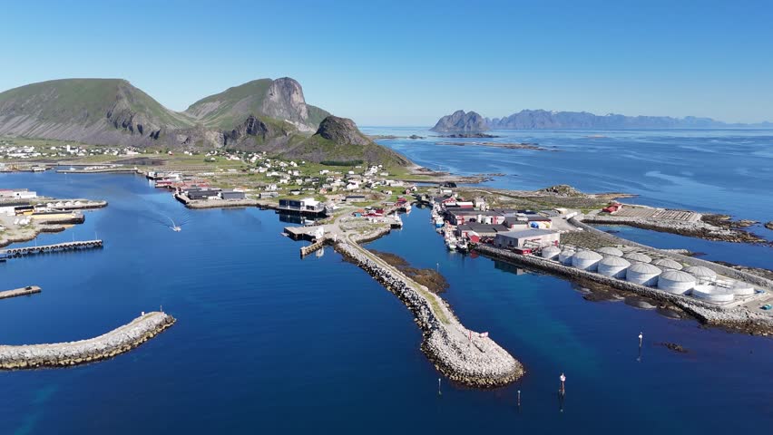 Aerial view of Værøy harbor: colorful village by the sea, dramatic mountains rising behind, and the endless blue of the Norwegian coast.