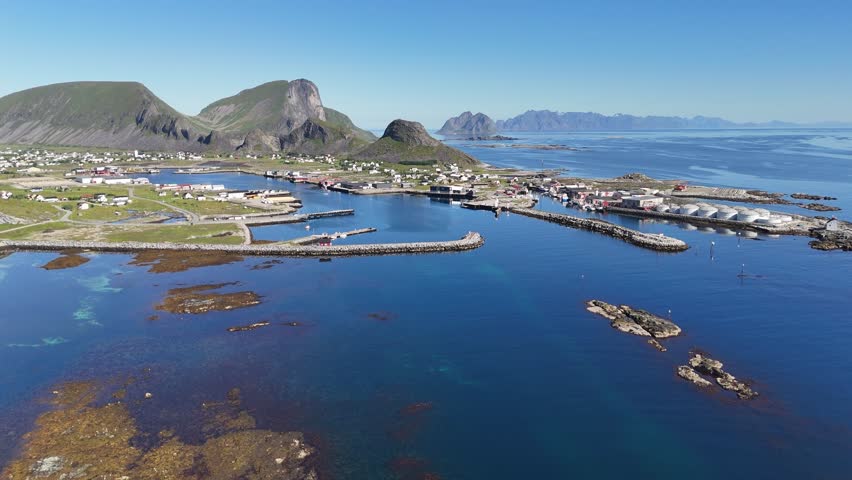 Aerial view of Værøy harbor: colorful village by the sea, dramatic mountains rising behind, and the endless blue of the Norwegian coast.