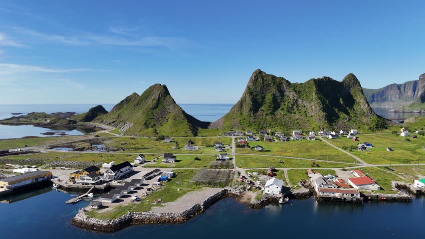 Aerial view of Værøy harbor: colorful village by the sea, dramatic mountains rising behind, and the endless blue of the Norwegian coast.