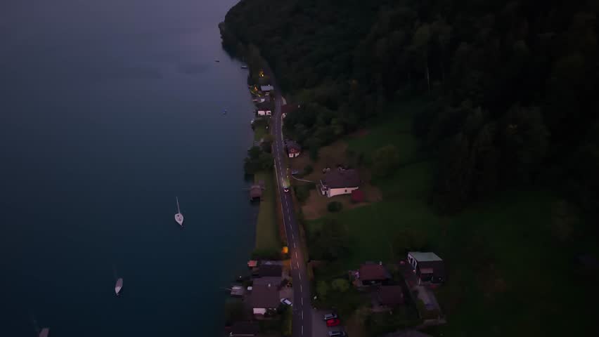 An elevated drone view at dusk of a lakeside village road lined with cozy homes, boats floating on still waters nearby