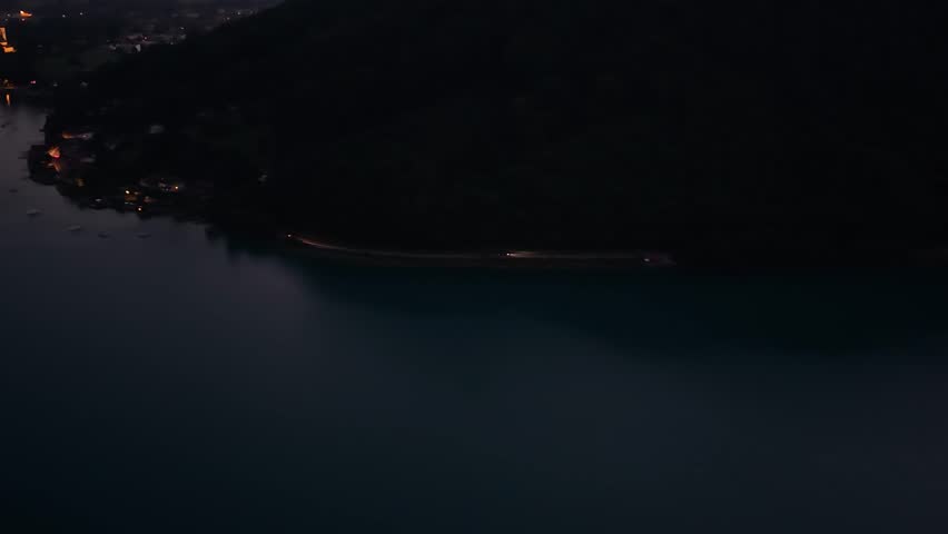 An aerial night view of a winding lakeside road curving between dark forested hills and calm water under fading light