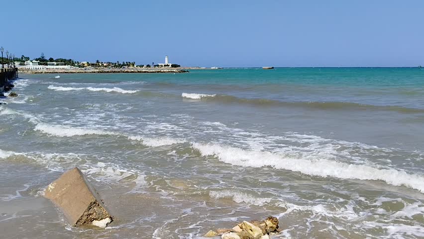 Gentle waves roll onto a shoreline with a large concrete block as a white lighthouse stands in the distance under a clear blue sky.