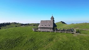 Aerial view of the wooden chapel surrounded by a fence on a green meadow with scattered rocks and wooden houses, under the backdrop of snow-capped mountains, Velika Planina, Kamnik, Slovenia. - Powered by Shutterstock - Get 15% off with code: PIKWIZARD15