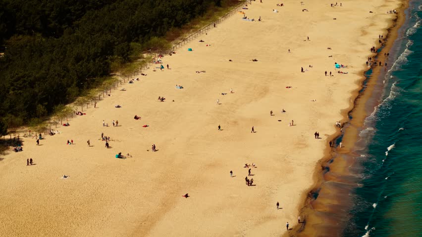 Sandy coastline with gentle waves and visitors in Gdansk Poland. People walking and relaxing along golden beach. Forest edge lining tranquil seaside environment. Natural beauty of Baltic shore under