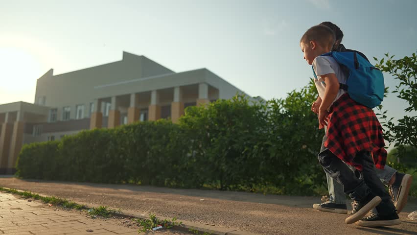 Boy and girl holding hands walk slowly on paved path to school. Morning light shines over backpacks. Student children follow daily school walk. Boy walks path to school with child sibling beside him