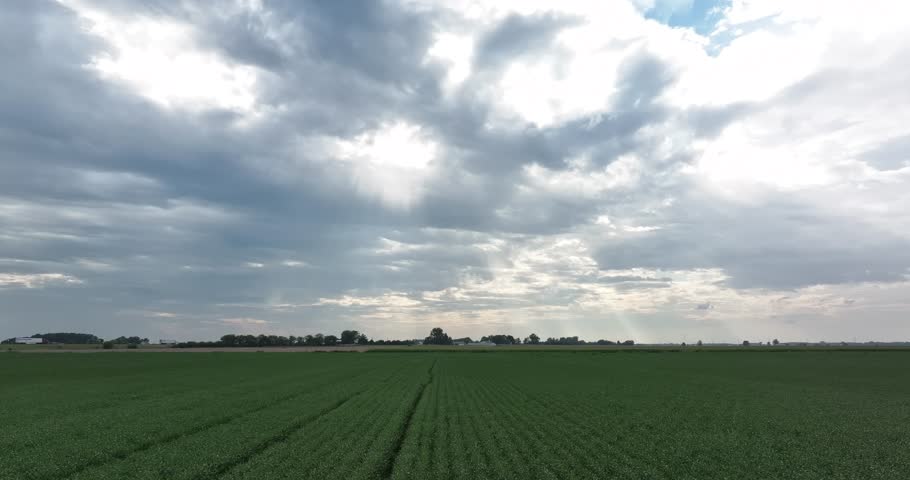 Aerial view of a vast green field stretches towards the horizon under a dramatic sky with sun rays piercing through the clouds, Monroeville, Indiana, United States.