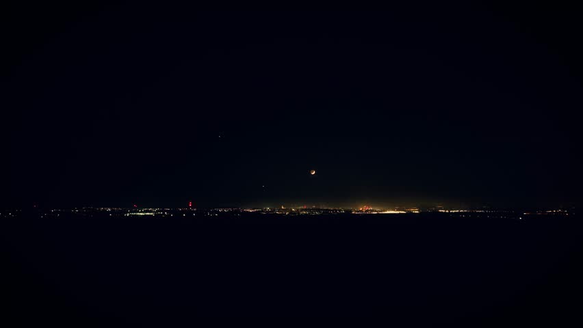 Crescent moon glowing above lights of night city. Lunar shape shining over illuminated urban skyline. Thin moon rising quietly in dark sky over glowing buildings. Nightscape sparkling under soft