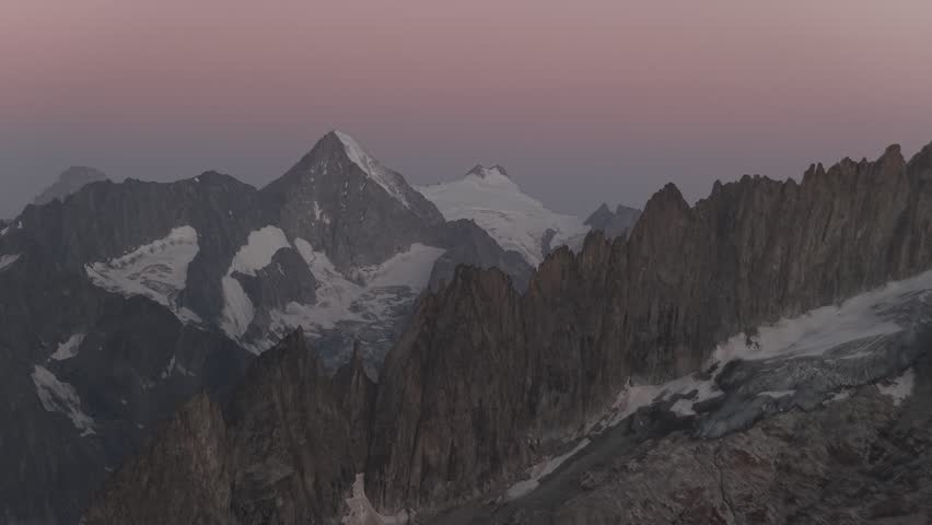 Aerial view of jagged, snow-capped mountains under a dusky sky, their peaks piercing the horizon with stark beauty, Fieschertal, Valais, Switzerland.