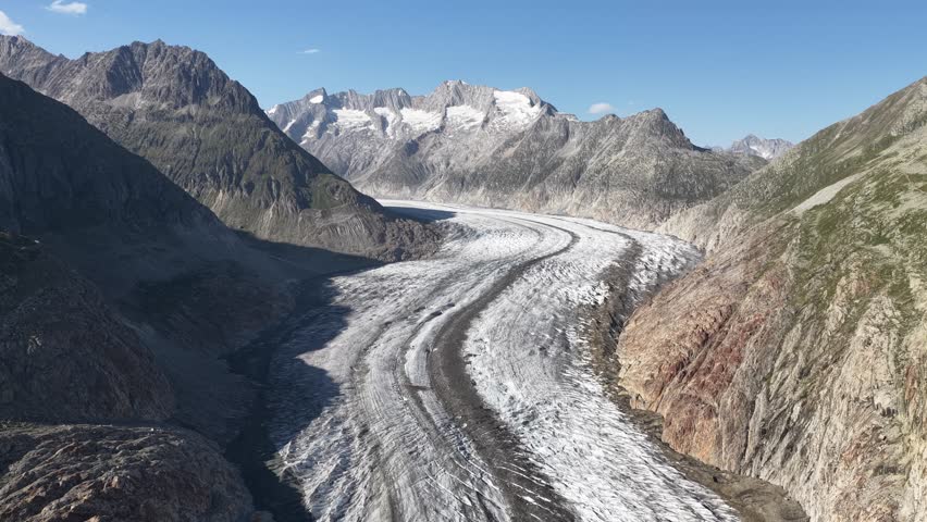 Aerial view of the Aletsch Glacier winding through rocky mountains under a clear blue sky, a striking contrast of ice and stone, Fieschertal, Valais, Switzerland.