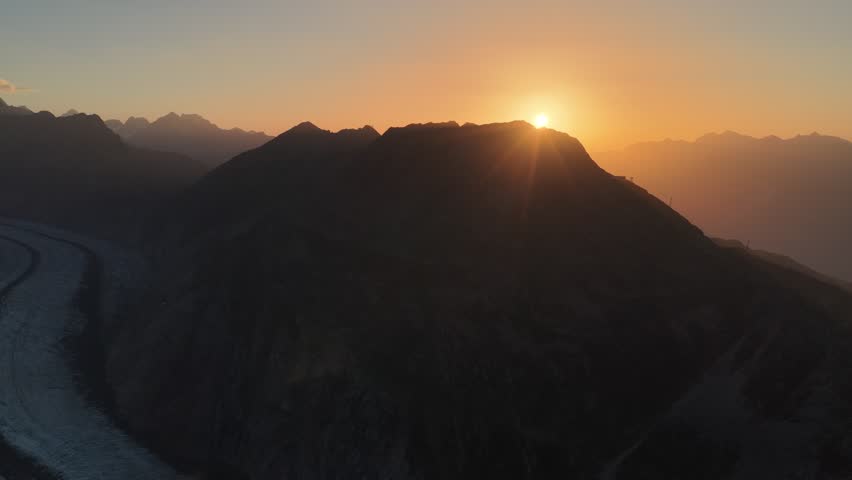Aerial view of a glacial river winding through a dramatic mountain range at sunset, showcasing contrasting tones and textures, Fieschertal, Valais, Switzerland.