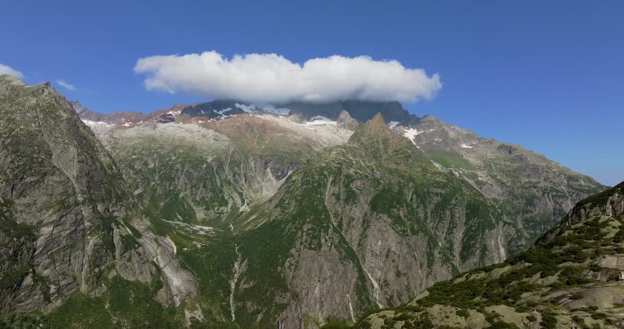 Alpine landscape featuring towering granite peaks and lush green slopes under a vivid blue sky, thick cloud formation across the mountaintops