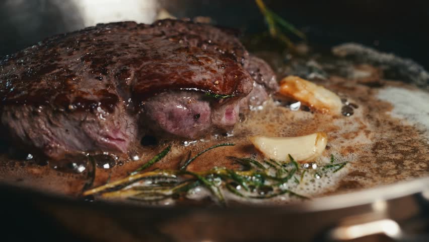 A closeup footage of a steak being seared in a pan with melted butter, garlic cloves, and fresh rosemary sprigs, with blurred background