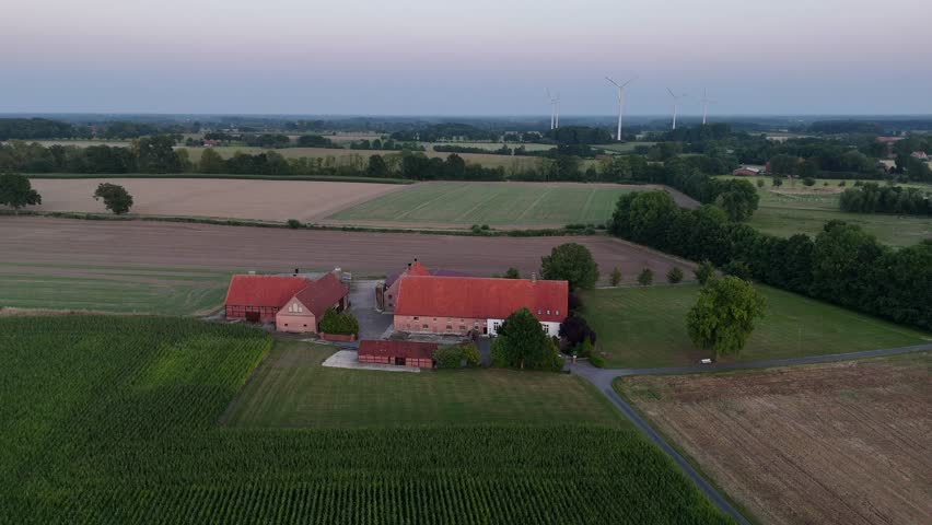 Traditional German farmhouse with red roofs, surrounded by fields, trees and wind turbines in rural countryside at sunset. Aerial orbiting shot. Dusk scene in Germany. Large property area in suburb.
