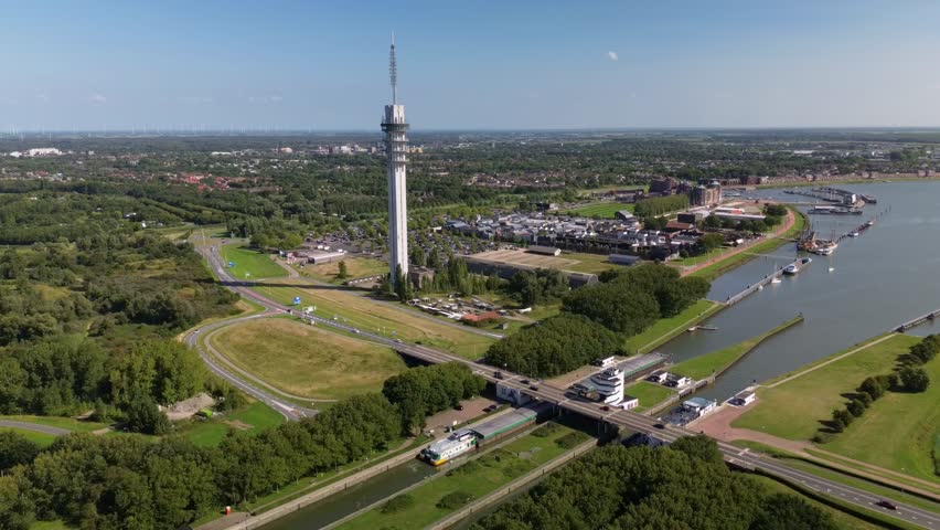 A tall communication tower rises above a riverside city with canals, bridges, boats, and green landscapes, blending modern infrastructure with natural surroundings.