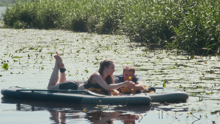 Wide shot of young Caucasian man giving flower to girlfriend and kissing while lying on paddle boards on river during outdoor date
