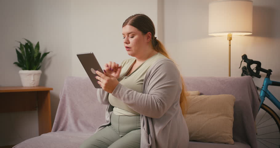 Overweight woman holds tablet and scrolls news feed looking focused. Woman with long hair tilts head slightly reading digital content sitting on sofa