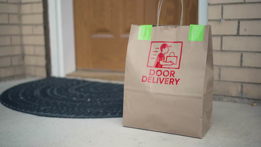 A homeowner retrieves a brown paper bag labeled “Door Delivery” from the front doorstep. The food delivery bag sits against the entry door, representing modern convenience and contactless service.