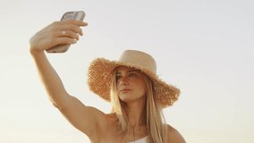 Young woman enjoys golden hour at beach while taking a selfie, wearing a stylish straw hat, capturing the moment filled with joy and relaxation - Powered by Shutterstock - Get 15% off with code: PIKWIZARD15