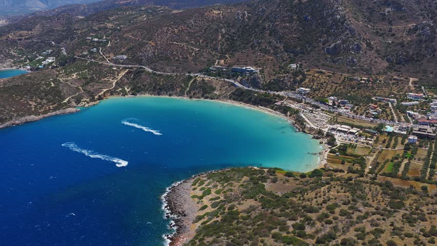 Aerial view of Voulisma Beach in Crete, showing turquoise to deep blue waters, rugged hills, winding roads, and boats leaving white trails in motion.
