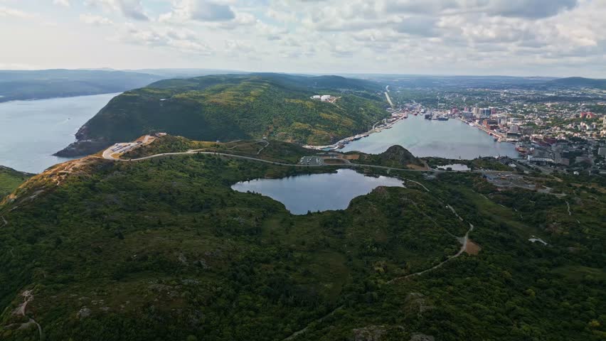 An aerial panorama captures Signal Hill’s rugged cliffs and lush greenery, with historic Cabot Tower on the headland and St. John’s harbour and city skyline stretching below.
