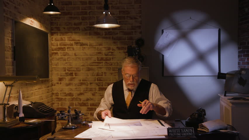 A scientist or engineer in 1945-1947 reaches for a model of the V2 rocket and ponders it as he studies some blueprints and other information on his desk.